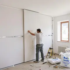 Un ouvrier installe des panneaux de placo pour une future cuisine. Des sacs de matériaux sont au sol.