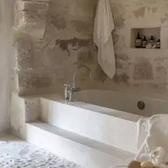 Bain relaxant dans une **salle de bain méditerranéenne** aux murs de pierre brute. Serviette blanche suspendue, robinetterie moderne.