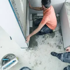 Un ouvrier réalise l'installation d'une douche, posant des panneaux muraux. Outils et matériaux de construction sont visibles au sol.