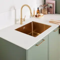 Évier en cuivre sous un robinet doré, sur un plan de travail stratifié blanc brillant. Des placards verts complètent l'ensemble.