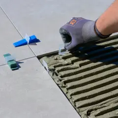 Pose de carreaux : un gant ajuste un coin pour enlever colle carrelage sèche. Des cales vertes et bleues sont déjà en place.