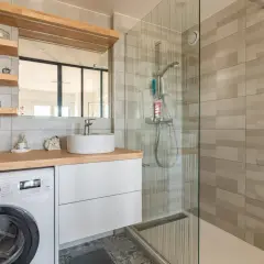 Coin buanderie dans salle de bain moderne avec lave-linge, lavabo sur meuble blanc et douche vitrée.