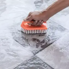 Main tenant nettoie une tache acide sur carrelage avec une brosse orange.
