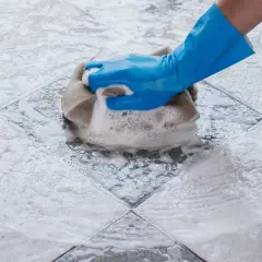 Main bleue frottant un carrelage avec une éponge, créant un voile de ciment savonneux.