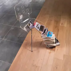 Une barre de seuil discrète sépare un carrelage sombre d'un parquet clair. Une chaise transparente et des livres créent une scène artistique.