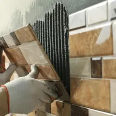 Mains gantées en train d'enlever du carrelage mural, révélant le mortier frais appliqué.