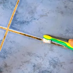 Une main utilise une brosse à dents pour blanchir le joint de carrelage sale, révélant la propreté cachée.