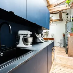 Cuisine moderne avec armoires bleues, évier noir et robots culinaires. L'espace offre une entrée directe dans la cuisine, avec des poutres apparentes et des plantes vertes.