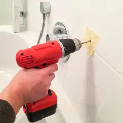 Une main tient une perceuse rouge pour percer du carrelage blanc, avec du ruban adhésif pour guider le foret.