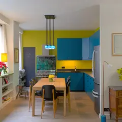 Plan cuisine ouverte sur salle à manger. Meubles bleus vifs, murs jaune citron, table en bois, chaises noires.