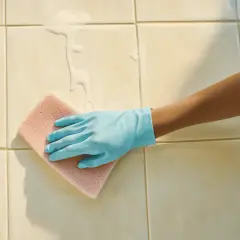 Main tenant, une main gantée de bleu frotte le carrelage de la salle de bain avec une éponge rose, faisant mousser le produit nettoyant.