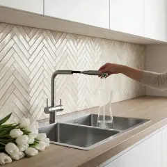 Une femme remplit un verre d'eau dans un évier de cuisine moderne. Lequel est la meilleure matière pour un évier de cuisine ? Le métal semble pratique.