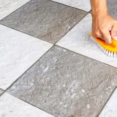 Main tenant, une brosse jaune frotte un carrelage blanc qui devient gris, révélant sa saleté.