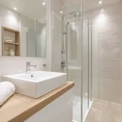 Une petite salle de bain moderne avec une douche vitrée, un lavabo rectangulaire sur un meuble en bois clair, et une serviette blanche pliée.