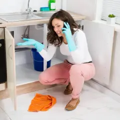 Une femme en gants bleus, au téléphone, tente de colmater une fuite d'eau sous son évier, un seau bleu à ses pieds.