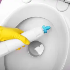 Main jaune appliquant un produit pour éliminer le calcaire dans une toilette blanche.