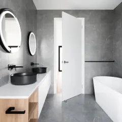 Salle de bain moderne aux murs en **calcaire** gris, avec double vasque noire, baignoire îlot et miroirs lumineux.