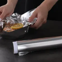 Mains de femme manipulant un saumon cuit dans du papier aluminium, prêt pour le nettoyage. Le rouleau d'aluminium est à côté.