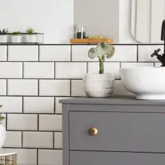 Planifier la dimension vasque salle de bain : un meuble gris, une vasque blanche, un robinet noir et des plantes vertes sur fond de carrelage blanc.