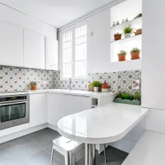 Cuisine moderne aux meubles blancs épurés, rehaussée par une crédence aux motifs géométriques gris et blancs. Une table blanche intégrée et des plantes apportent une touche de fraîcheur.