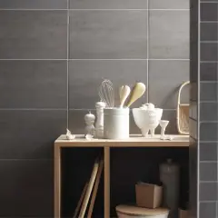 Sur un mur en carrelage gris, des ustensiles de cuisine et des objets décoratifs sont disposés sur une étagère en bois. La faïence du mur de droite est plus petite.