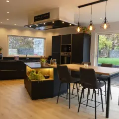 Cuisine moderne avec îlot central, plan de travail noir et carrelage marron foncé. Table haute avec tabourets, luminaire design.