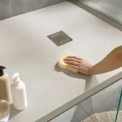 Main d'une personne nettoyant un receveur de douche à éviter avec une éponge. Panier de serviettes et bouteilles de savon à proximité.