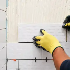 Mains gantées jaunes en pleine pose carrelage mural blanc. Des croisillons maintiennent l'espacement.