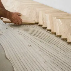 Mains d'un artisan en train de poser du parquet en chevrons sur un lit de colle ondulée.