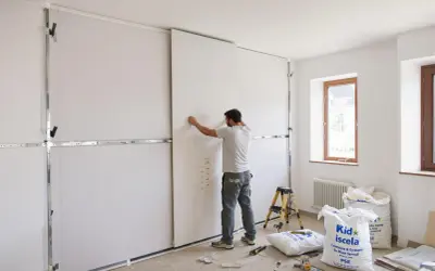 Un ouvrier installe des panneaux de placo pour une future cuisine. Des sacs de matériaux sont au sol.