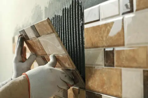 Mains gantées en train d'enlever du carrelage mural, révélant le mortier frais appliqué.