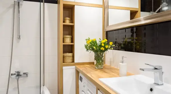 Petite salle de bain avec baignoire, lavabo, machine à laver et étagères en bois. Un bouquet de fleurs jaunes décore le plan de travail.