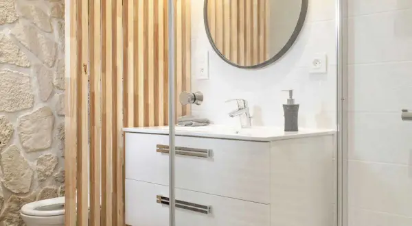 Salle de bain moderne avec un lavabo suspendu, un miroir rond et une **installation WC** discrète derrière un paravent en bois.