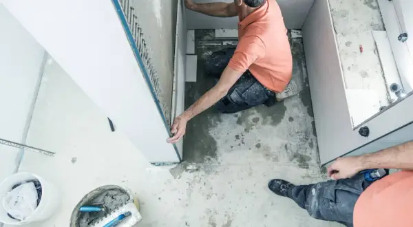 Un ouvrier réalise l'installation d'une douche, posant des panneaux muraux. Outils et matériaux de construction sont visibles au sol.