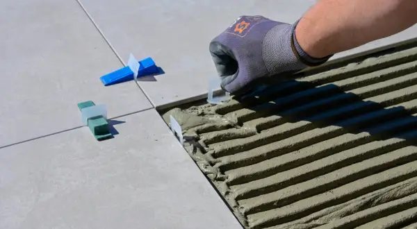 Pose de carreaux : un gant ajuste un coin pour enlever colle carrelage sèche. Des cales vertes et bleues sont déjà en place.