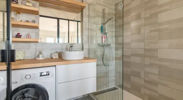 Coin buanderie dans salle de bain moderne avec lave-linge, lavabo sur meuble blanc et douche vitrée.