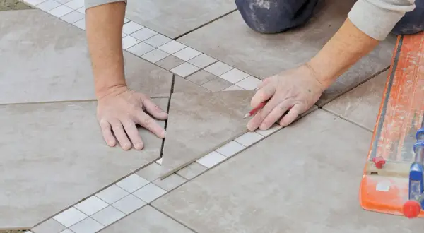 Pose en quinconce de carreaux de sol, avec une bordure décorative. Un carreleur marque une pièce avec un crayon.