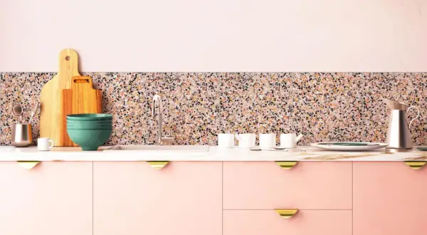 Cuisine moderne avec des armoires roses, un plan de travail en marbre et un crédence en terrazzo coloré. Ustensiles de cuisine et vaisselle disposés.