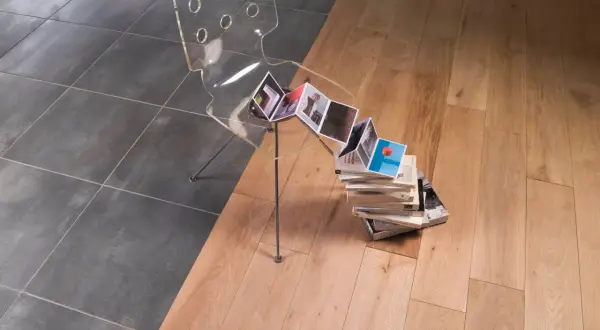 Une barre de seuil discrète sépare un carrelage sombre d'un parquet clair. Une chaise transparente et des livres créent une scène artistique.