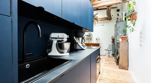 Cuisine moderne avec armoires bleues, évier noir et robots culinaires. L'espace offre une entrée directe dans la cuisine, avec des poutres apparentes et des plantes vertes.