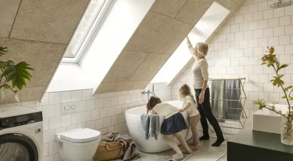 Salle de bain sous pente avec wc suspendu. Une femme et deux enfants admirent la lumière entrant par la fenêtre de toit.