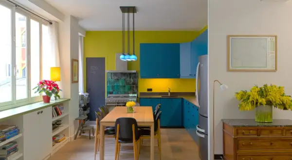 Plan cuisine ouverte sur salle à manger. Meubles bleus vifs, murs jaune citron, table en bois, chaises noires.