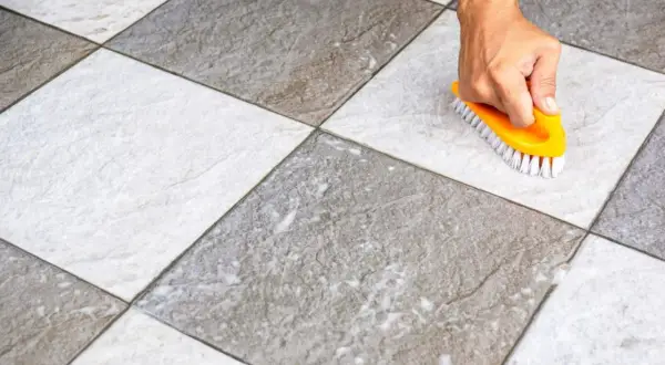Main tenant, une brosse jaune frotte un carrelage blanc qui devient gris, révélant sa saleté.