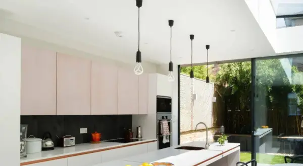 Cinq suspensions noires au design épuré, quel luminaire pour cuisine moderne ! Elles éclairent une cuisine lumineuse avec ses meubles roses et sa grande baie vitrée.
