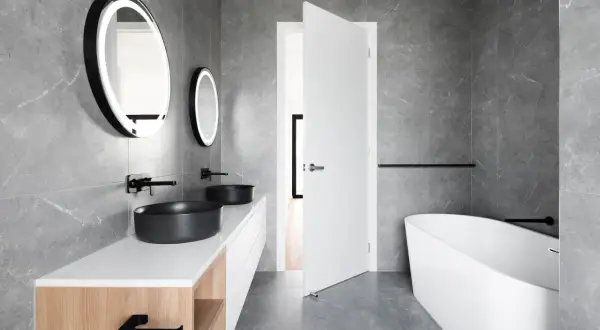 Salle de bain moderne aux murs en **calcaire** gris, avec double vasque noire, baignoire îlot et miroirs lumineux.