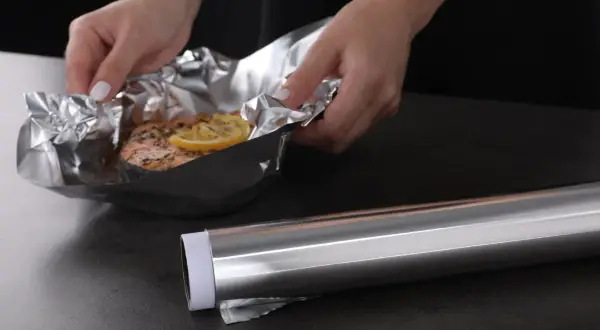 Mains de femme manipulant un saumon cuit dans du papier aluminium, prêt pour le nettoyage. Le rouleau d'aluminium est à côté.