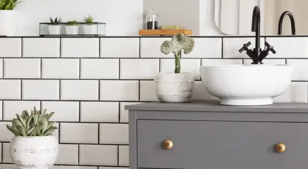 Planifier la dimension vasque salle de bain : un meuble gris, une vasque blanche, un robinet noir et des plantes vertes sur fond de carrelage blanc.