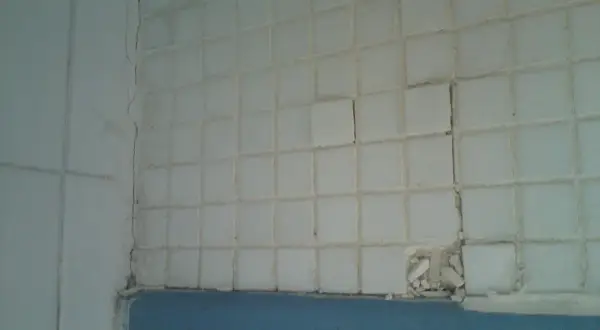 Carrelage blanc qui se décolle, craquelé et endommagé par l'humidité. Des carreaux sont cassés, révélant le mur derrière.