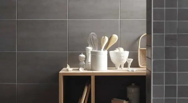 Sur un mur en carrelage gris, des ustensiles de cuisine et des objets décoratifs sont disposés sur une étagère en bois. La faïence du mur de droite est plus petite.