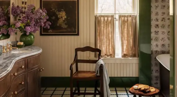 Ambiance de salle de bain rustique avec un lavabo en marbre, des lilas violets, un tableau ancien et une chaise en bois.